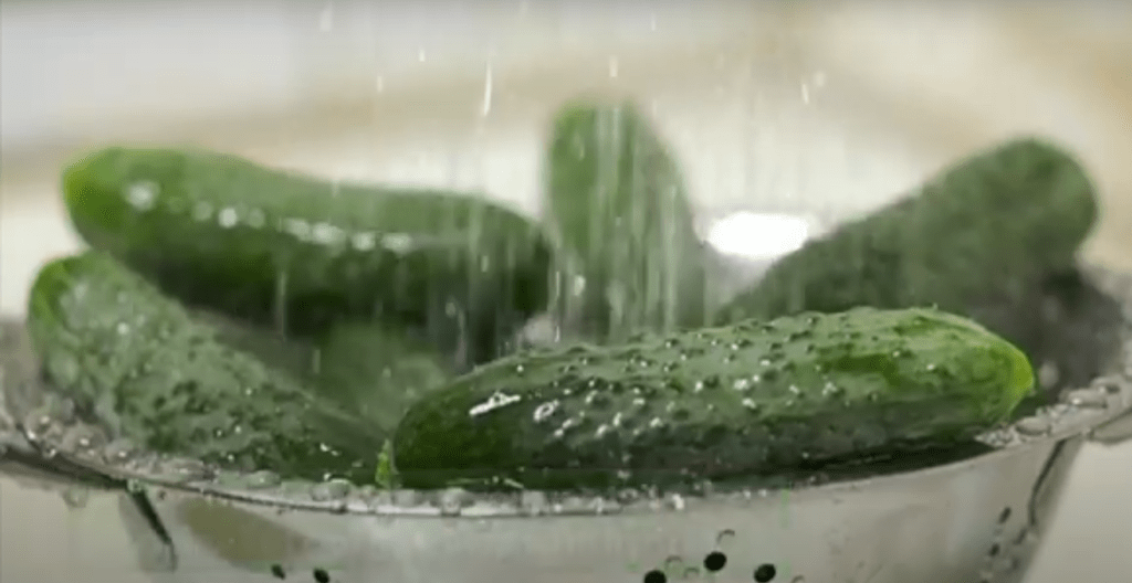 An overhead view of a bowl filled with fresh cucumbers, with a stream of water pouring over them. The water cascades down, creating droplets and a glistening effect on the cucumbers, emphasizing their freshness and cleanliness